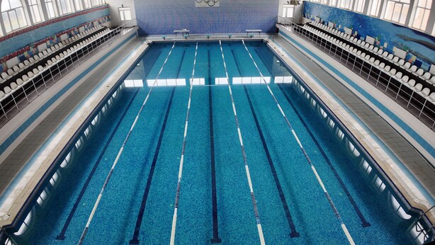 A large indoor swimming pool with multiple lanes, spectator seating, and a tiled wall with a mosaic design.