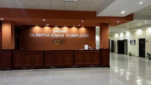 A reception area with a wooden desk, a sign reading "ŞAGAN OLMİPIYA İDMAN KOMPLEKSİ" and the Olympic rings logo. - Arena Hotel Shagan (Baku)