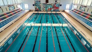 A large indoor swimming pool with multiple lanes, a diving board, and a spectator area with chairs.