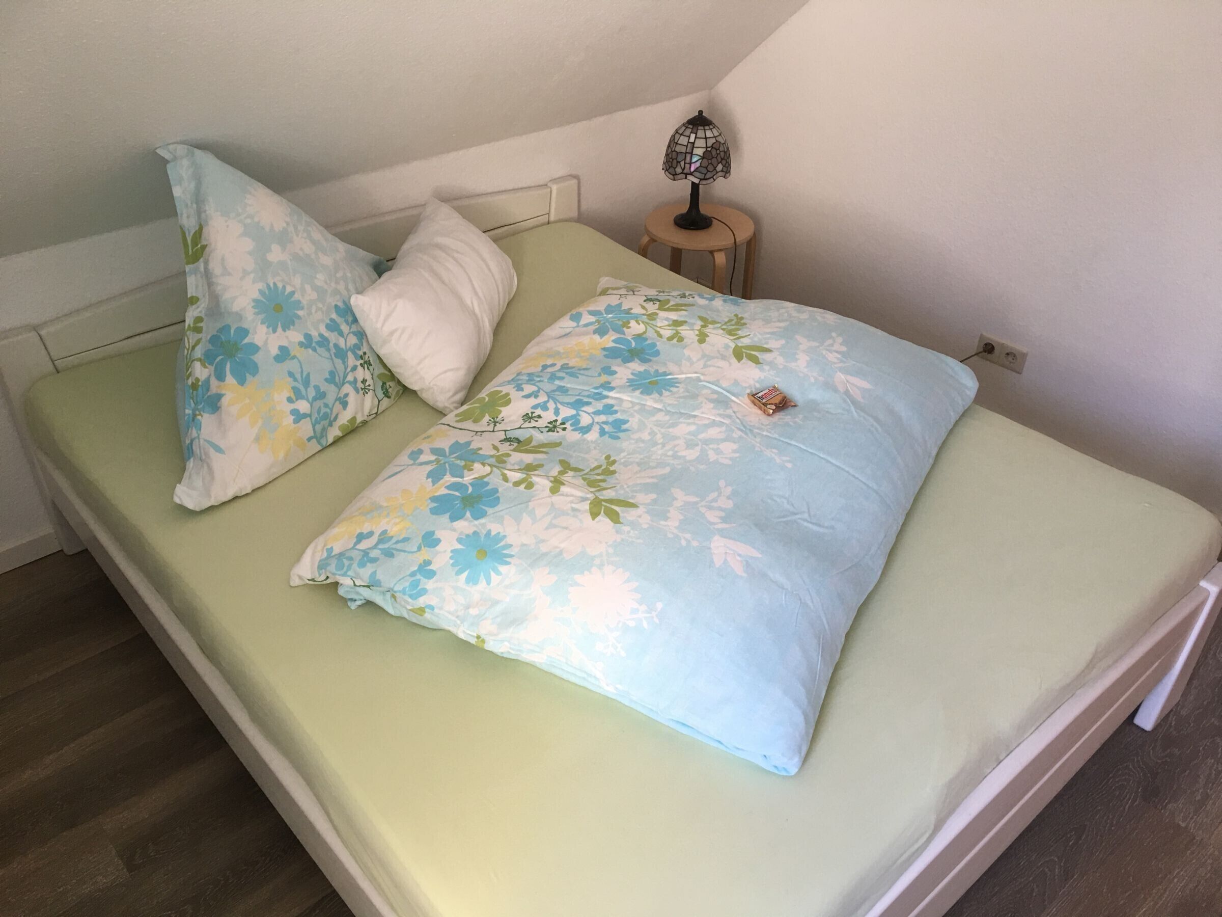 A single bed with floral bedding, a bedside lamp, and a power outlet on the wall.
