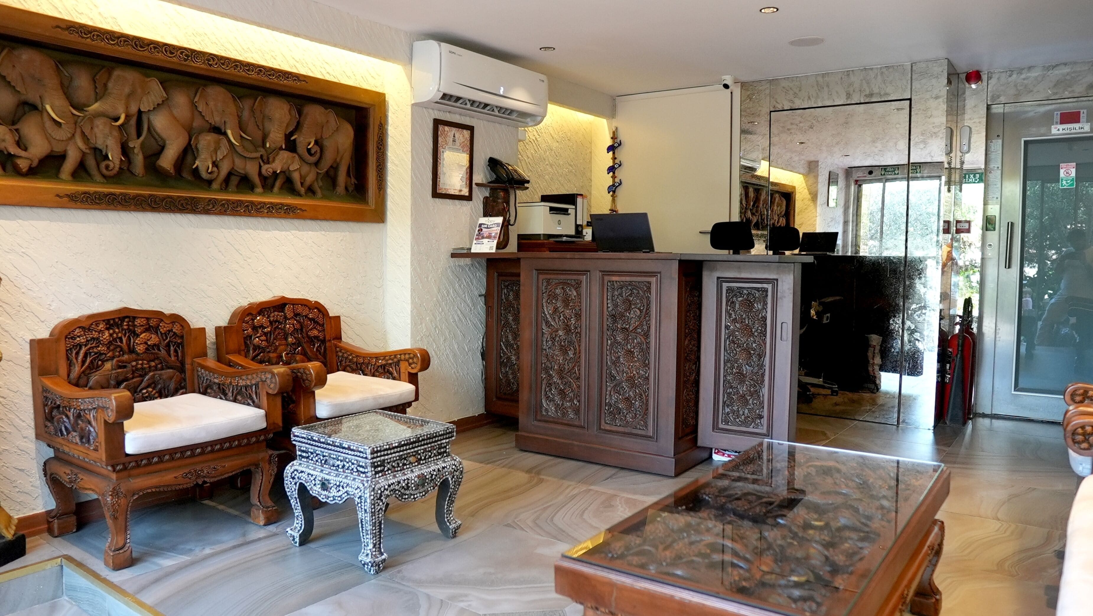 Un hall d’entrée agrémenté de fauteuils en bois sculpté, d’une table basse en verre et d’un comptoir de réception doté d’un téléphone et de tableaux encadrés.