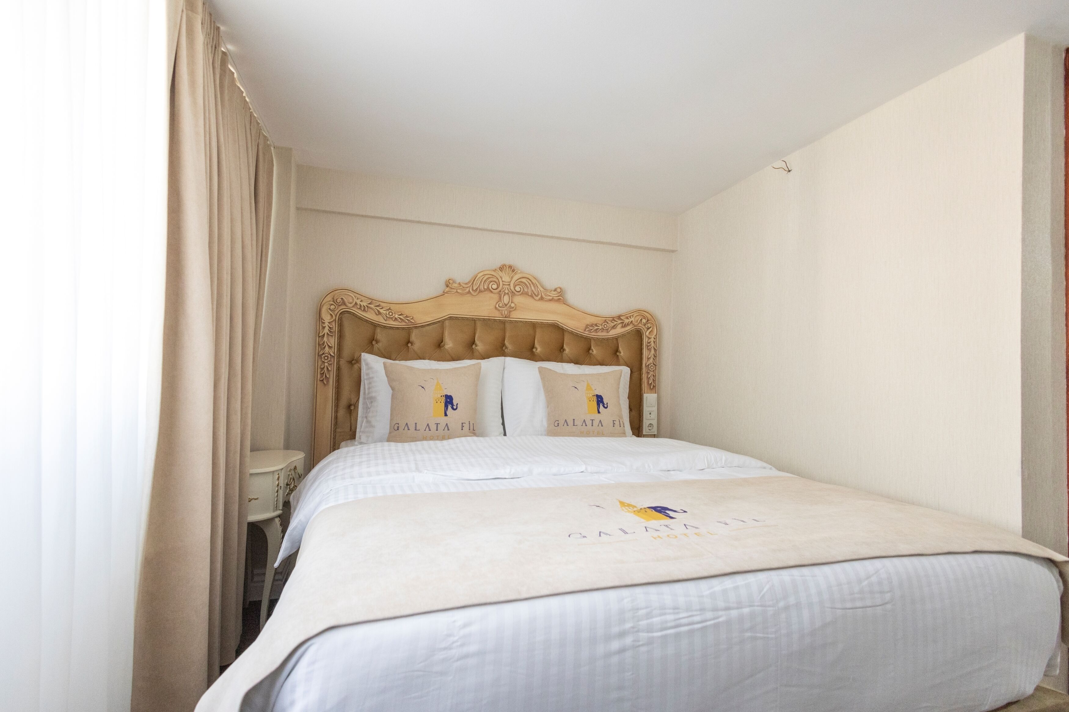 Habitación de hotel con una cama grande, un cabecero de madera, dos almohadas con el texto 'GALATA FLY' y una mesita de noche pequeña.
