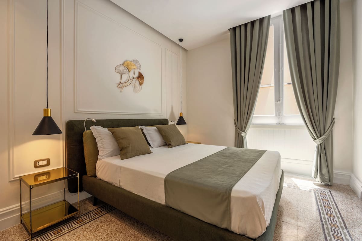 A neatly arranged bedroom with a bed, bedside lamps, a wall art piece, and a window with curtains.