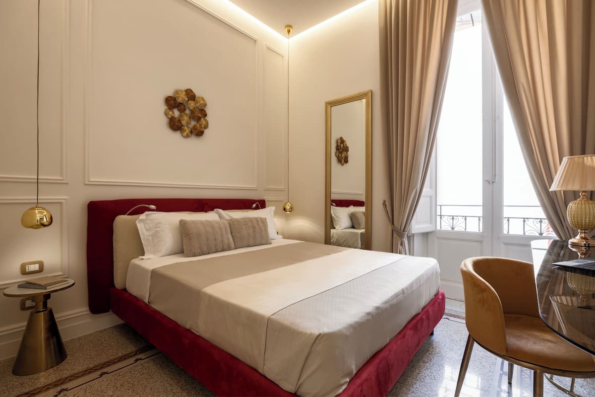 A hotel room with a large bed, a round table with a lamp, a chair, a small side table, and a mirror on the wall.