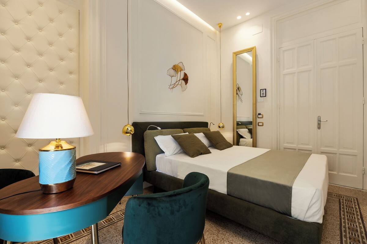 A hotel room with a tufted headboard, a bed with a green headboard, a blue armchair, a wooden side table with a lamp, and a mirror on the wall.