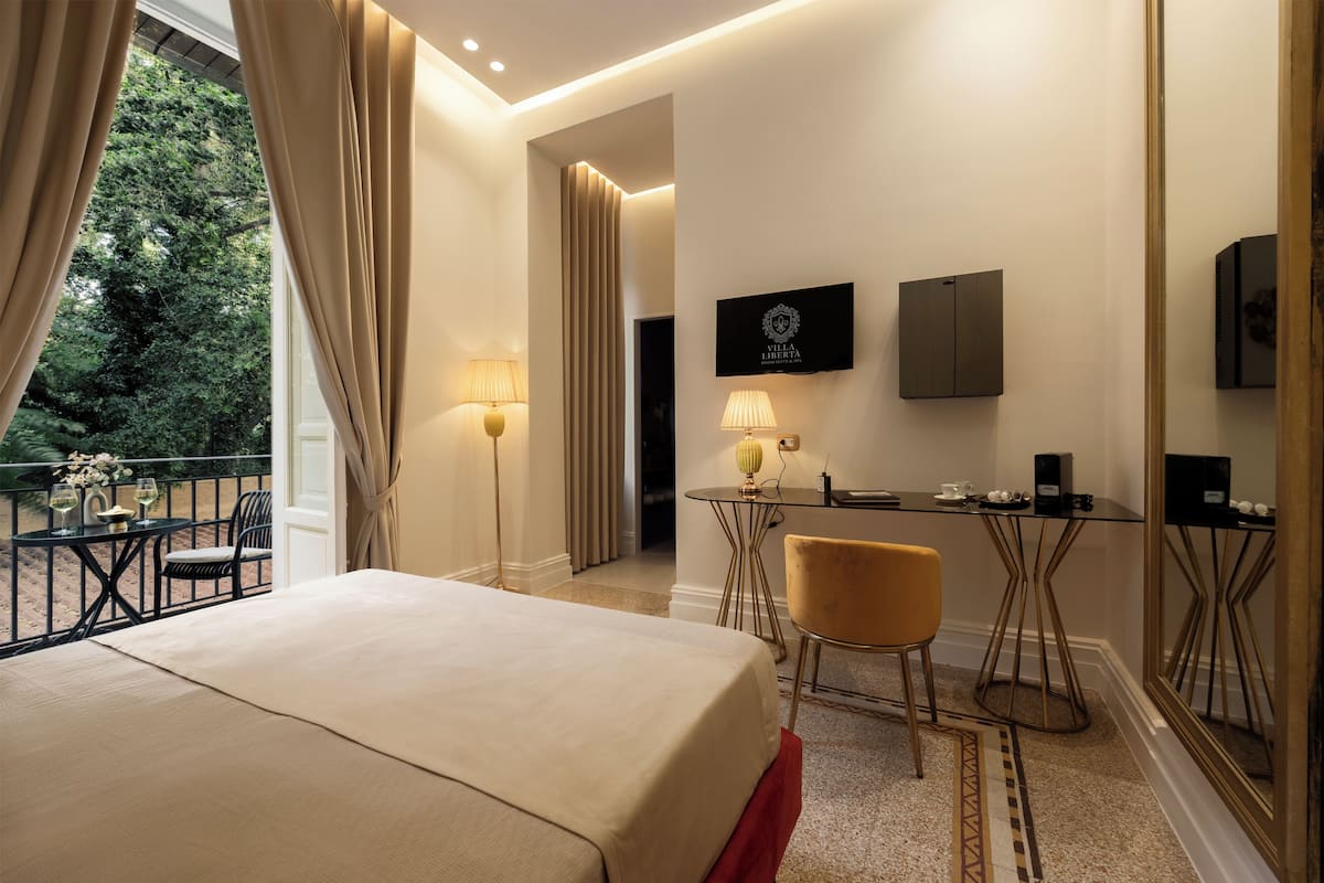 A hotel room with a bed, a desk with a chair, a TV, and a view of greenery outside.