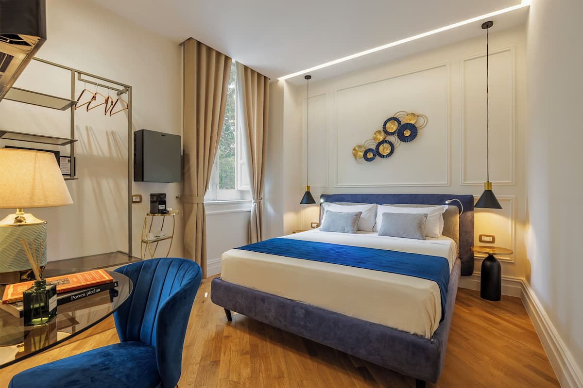A modern hotel room with a large bed, a seating area with a blue chair, a small table with books, and a decorative wall piece.
