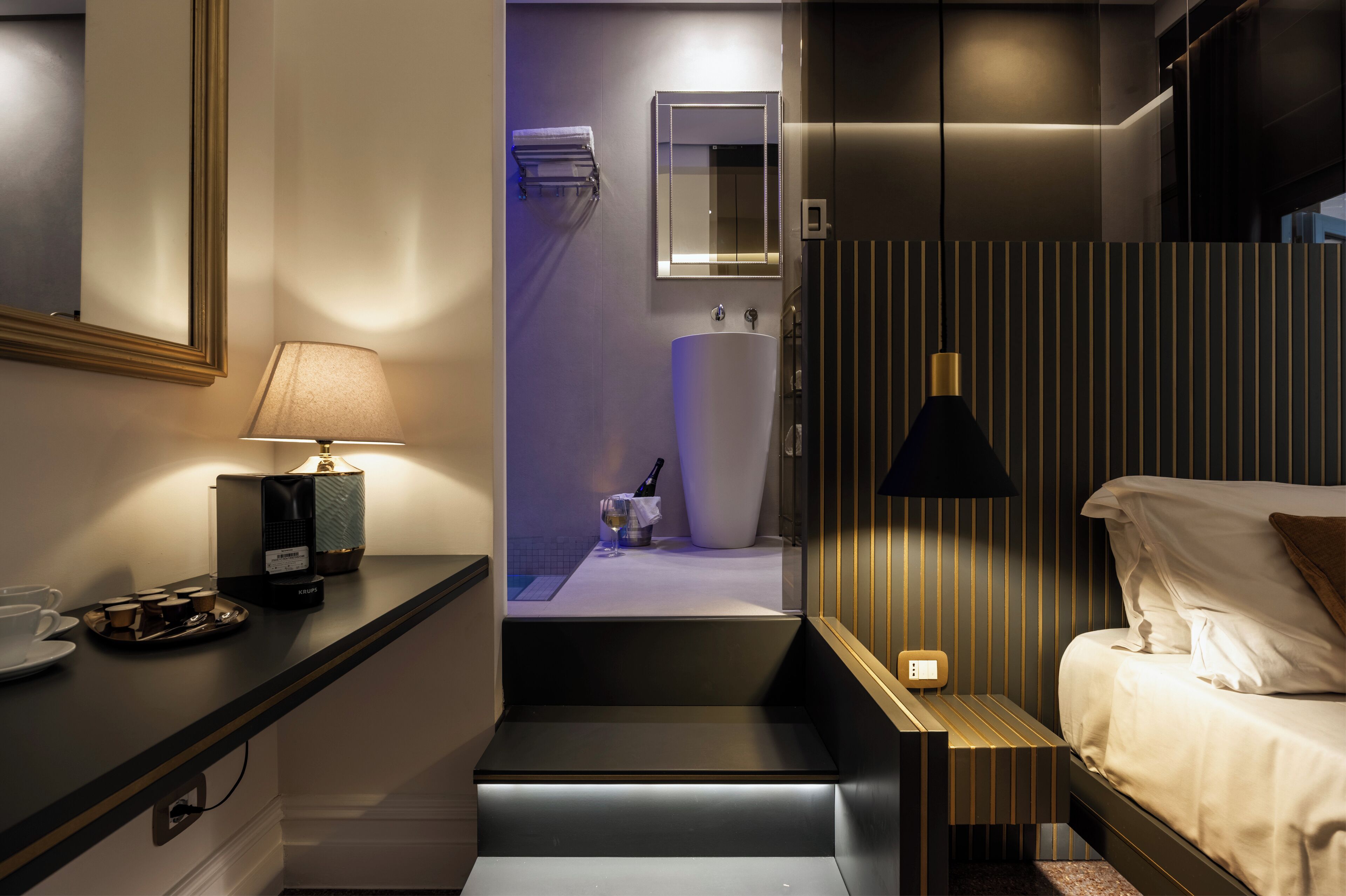 A modern hotel room with a bed, bedside table, lamp, and a small seating area with a tray of cups and a bottle.