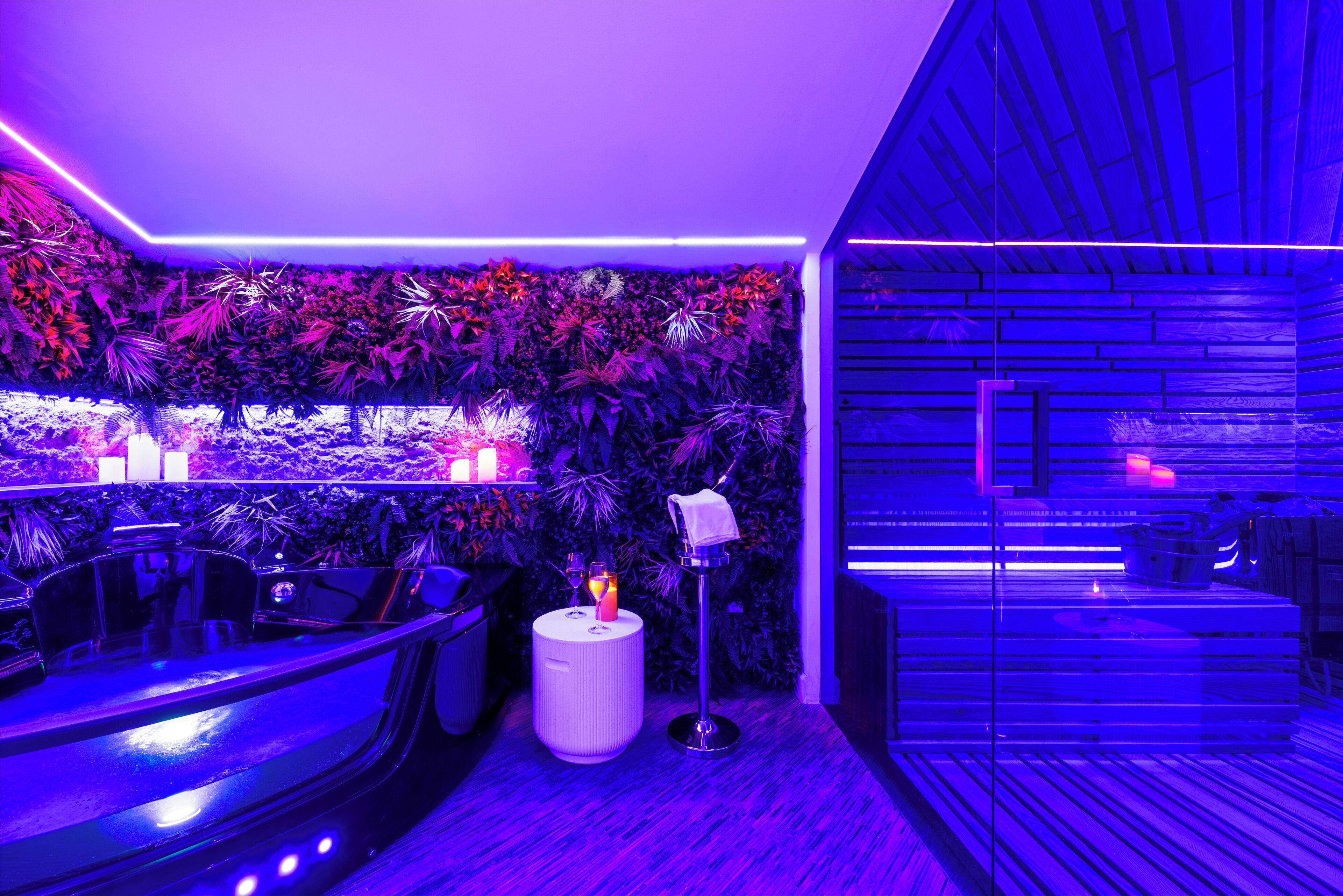 A modern bar area with a neon-lit ceiling, a seating area with a bar counter, and a wall adorned with lush green plants.