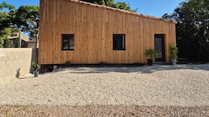 A wooden building with two windows and a door, surrounded by gravel and trees.