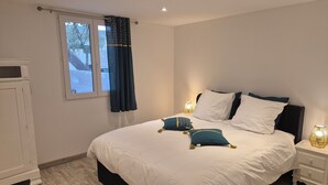 A bedroom with a large bed, white walls, a window with curtains, a bedside table with a lamp, and a nightstand.