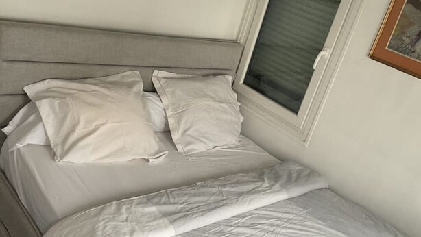 A bed with white bedding and a headboard, a window with blinds, and a framed picture on the wall. - Urban Shelter (Sarvelles)