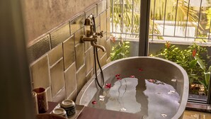 Una bañera con pétalos de rosa, un cabezal de ducha y una ventana con plantas.