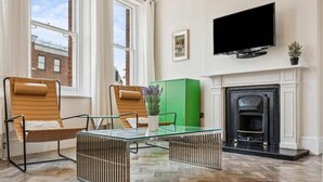 A living room with two leather chairs, a glass coffee table, a green cabinet, a fireplace, and a TV mounted on the wall. - Stunning 3-Bedroom Apartment in Hampstead (London)