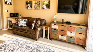 A living room with a brown leather sofa, a wooden side table, a TV mounted on the wall, and a wooden chest of drawers with numbered compartments.