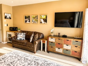 Ein Wohnzimmer mit einem braunen Ledersofa, einem Holznachttisch, einem an der Wand montierten Fernseher und einer Holzkommode mit nummerierten Fächern.