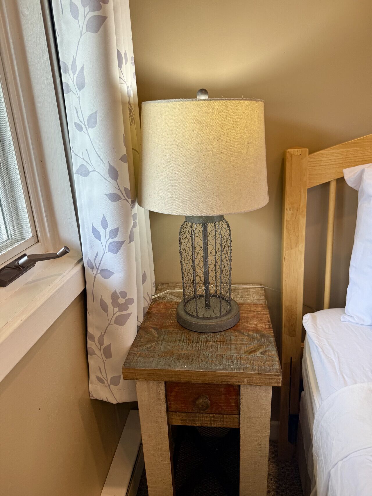 Una mesita de noche de madera con una lámpara, una cama y una ventana con cortinas.