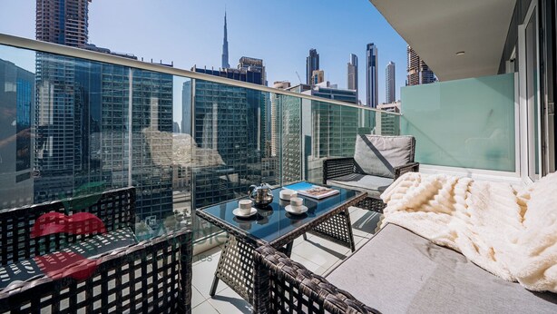 Un balcón con barandilla de vidrio, muebles de mimbre, una mesa con tetera y tazas, y vistas de un horizonte urbano con edificios altos.
