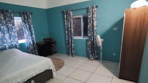 A bedroom with a bed, a desk, a chair, a window with curtains, and a door. - 2-bedroom apartment in lovely Sunny Acres with WiFi, AC and Fan (Choc)