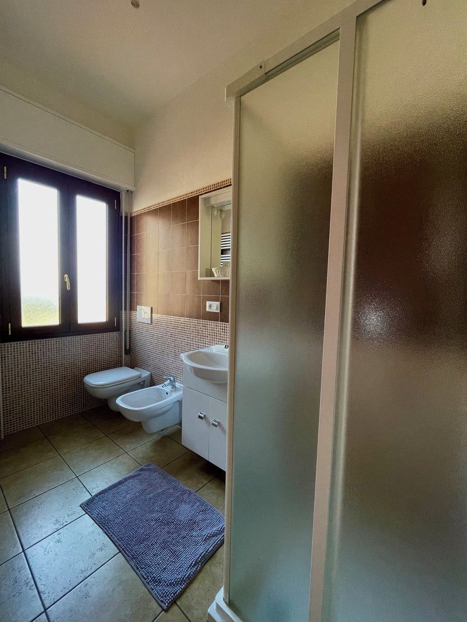 Une petite salle de bain comprenant des toilettes, un bidet et un lavabo. Une fenêtre laisse entrer la lumière naturelle et un miroir est placé au-dessus du lavabo.