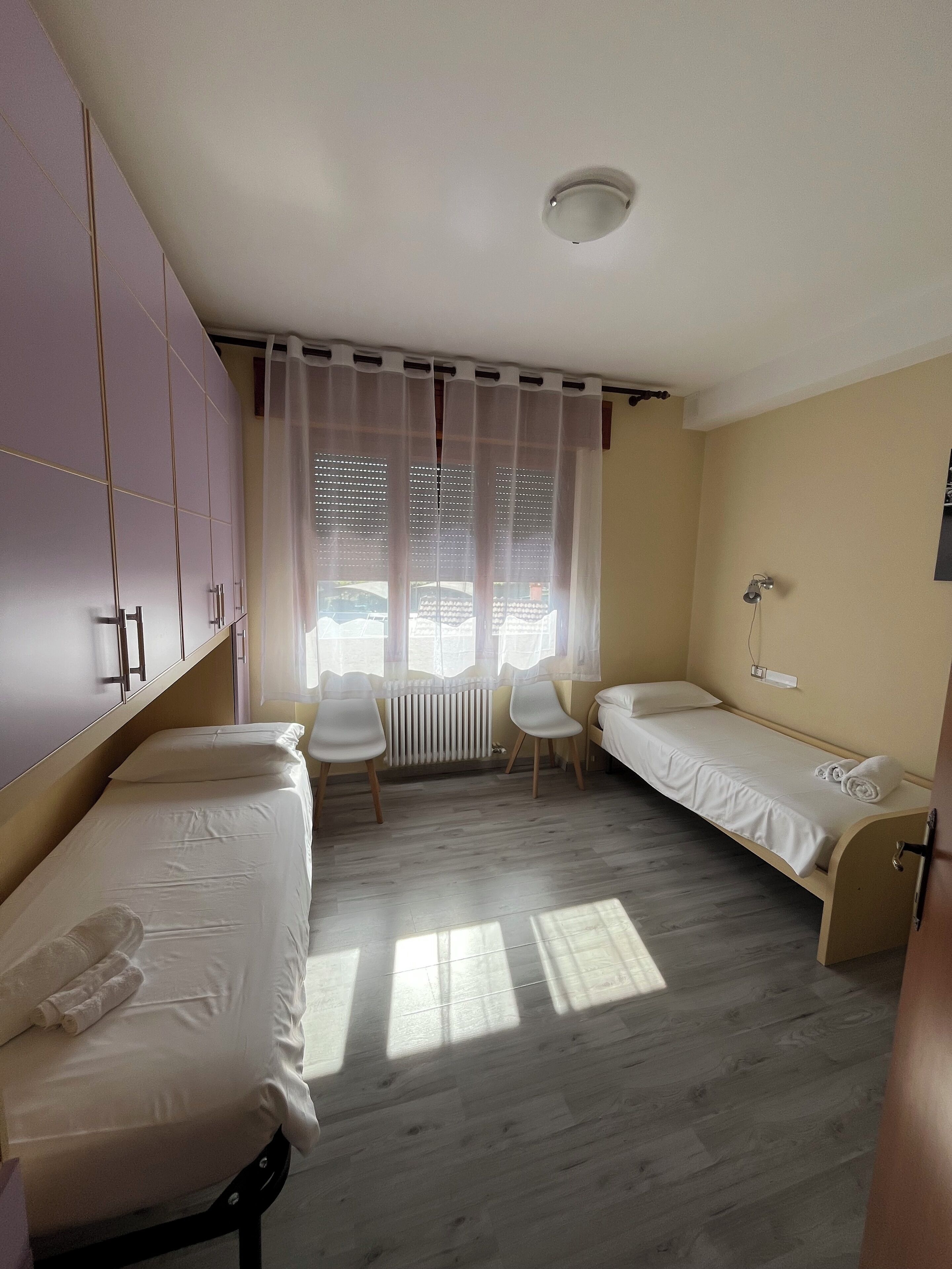 A room with two single beds, a chair, a radiator, and a window with sheer curtains.