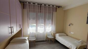 A room with two single beds, a chair, a radiator, and a window with sheer curtains.