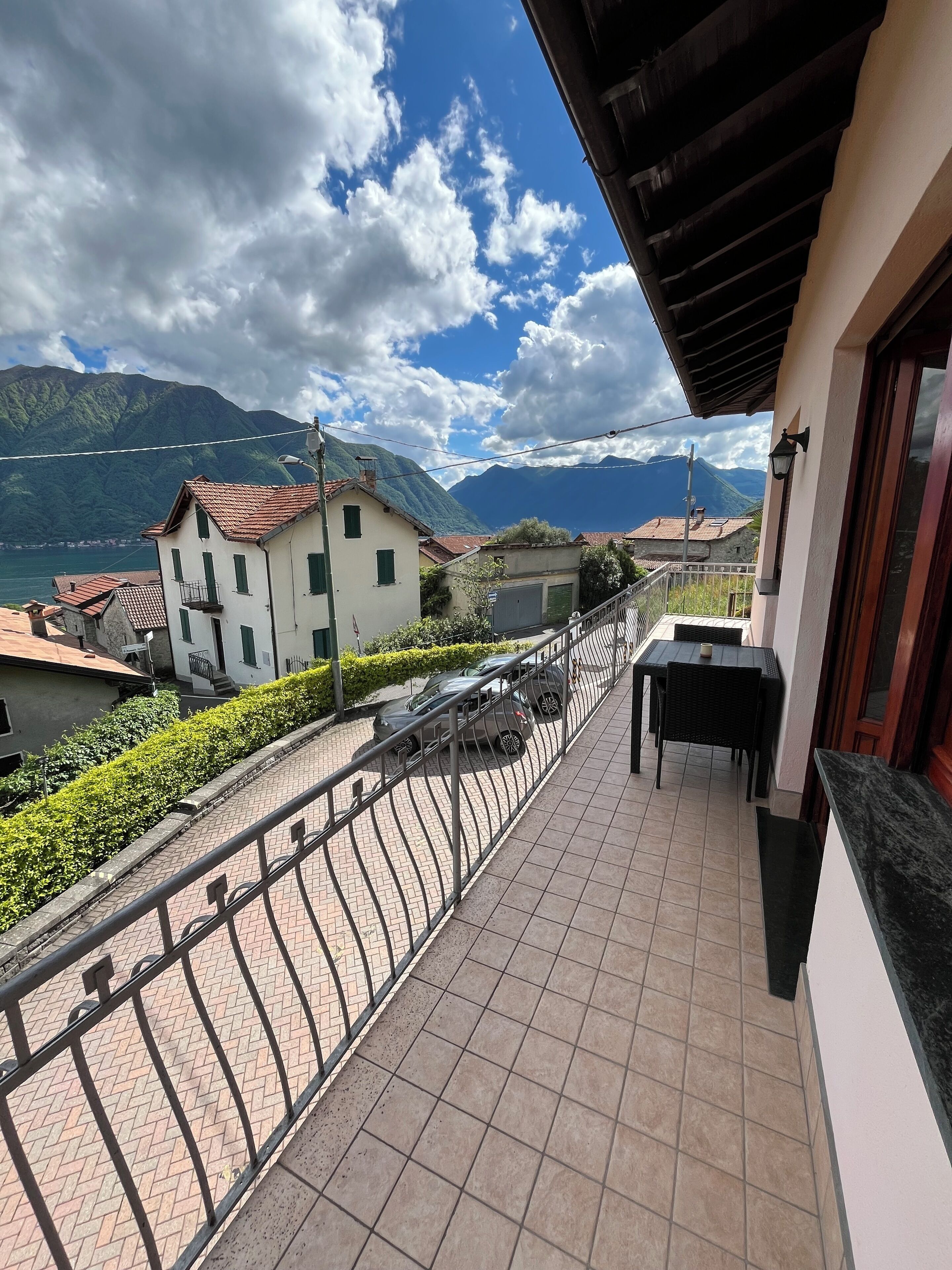A balcony with a tiled floor, a table, and chairs, overlooking a street with cars and buildings, with mountains in the background.