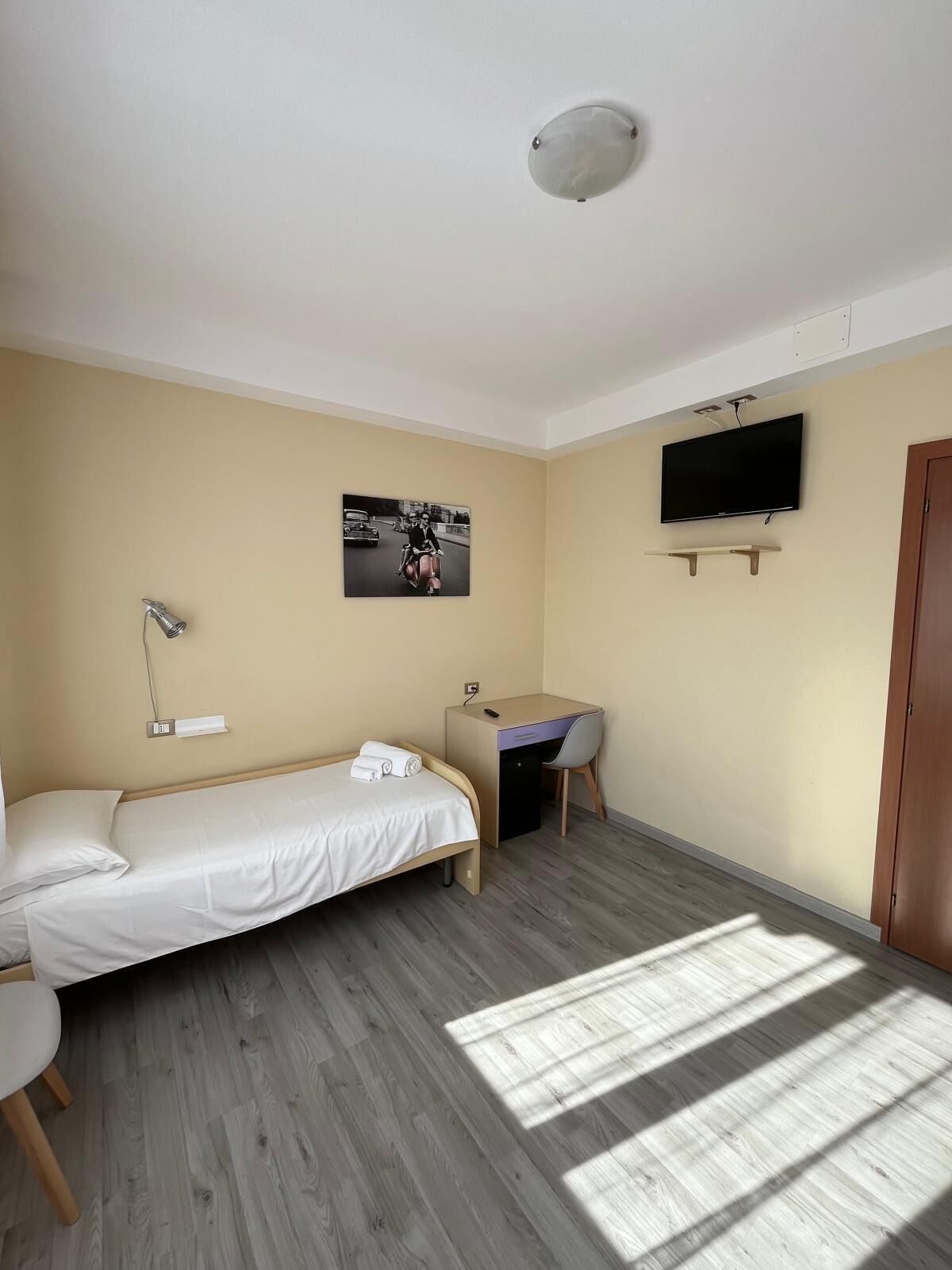 A small, well-lit room with a bed, a desk, a chair, a TV, and a picture on the wall.
