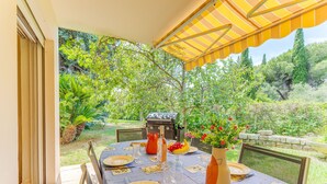 An outdoor dining area with a table set for two, a grill, and a view of the garden.