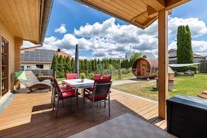 Eine Holzdiele mit Tisch und StĂŒhlen, eine Pergola und der Blick auf ein Haus mit Solaranlage.