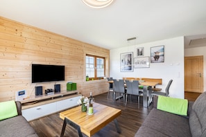 Un salon moderne doté d’un téléviseur à écran plat, de murs recouverts de panneaux en bois, d’un coin repas avec des chaises grises et d’un canapé agrémenté d’un coussin vert.