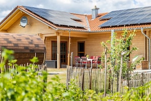 Une maison en bois avec un toit en tuiles, des panneaux solaires, un patio couvert et un jardin clôturé.