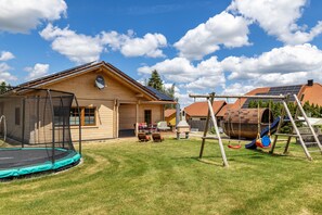 Une maison en bois avec un trampoline, une balançoire et une terrasse équipée de mobilier d’extérieur.