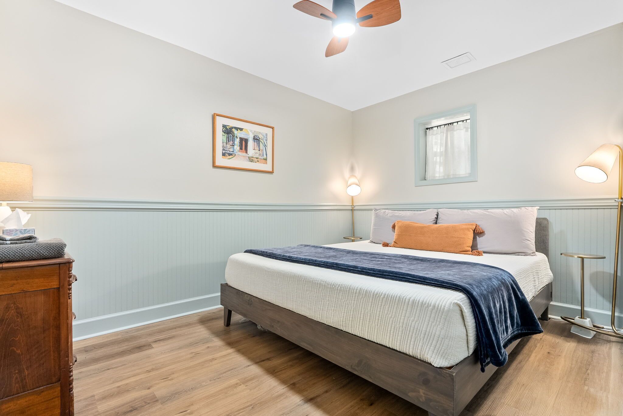 Une chambre à coucher avec un lit, une table de chevet en bois, un ventilateur de plafond, des lampes fixées au mur et un tableau encadré.