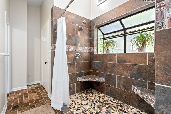 A shower area with a glass door, a window with plants, and a tiled floor.