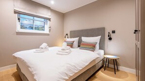 A neatly made bed with white linens and two pillows, a small bedside table with a lamp, and a window with a view of the outdoors. - F27 Living Whg. 01 - Sylt (Sylt)