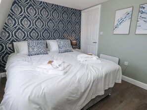 Ein ordentlich bezogenes Bett mit weißen Leinen und Handtüchern, Tapete mit Muster sowie zwei gerahmte Kunstwerke an der Wand.