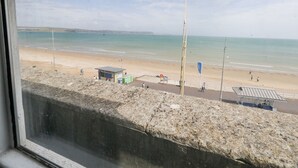 Uitzicht op een strand vanuit een raam, met zand, een badwachtershuisje, een vlag en enkele mensen.