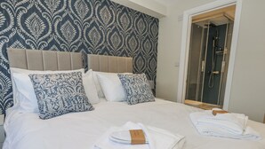 Un lit bien fait, avec du linge de lit blanc et des coussins décoratifs, une tête de lit en bois et une porte de douche en verre.