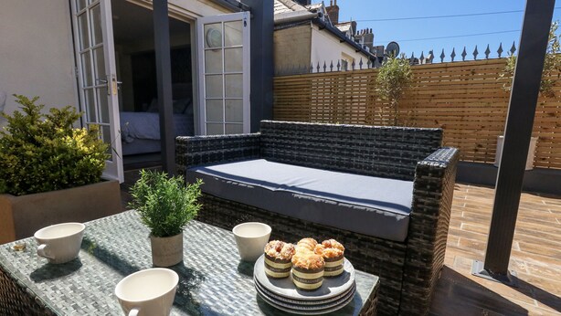 Une terrasse ensoleillée avec une table en osier, deux chaises, une petite plante en pot et un banc en osier agrémenté de coussins.