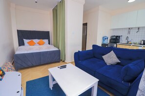 A compact hotel room with a bed, a blue sofa, a small table, and a kitchenette. - Pazoori Stays at Artesano Apartments (Syokimau)