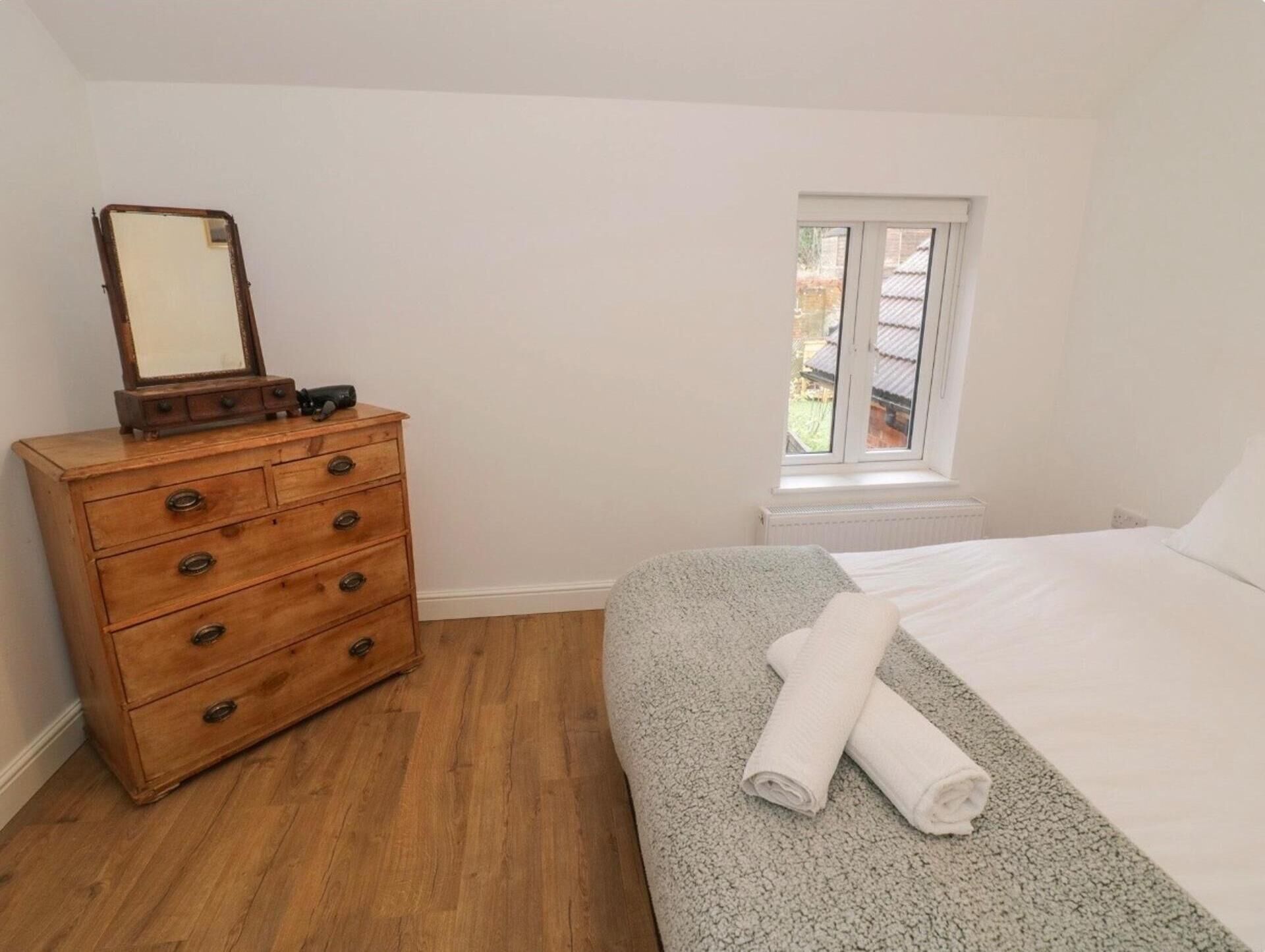 A bedroom with a wooden dresser, a bed with a grey blanket, and a window with a view of the outside.