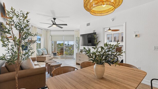 A spacious living room with a wooden dining table, a ceiling fan, and a large window overlooking a balcony. - August Beach Escape – Special Rates Available! (Treasure Island)