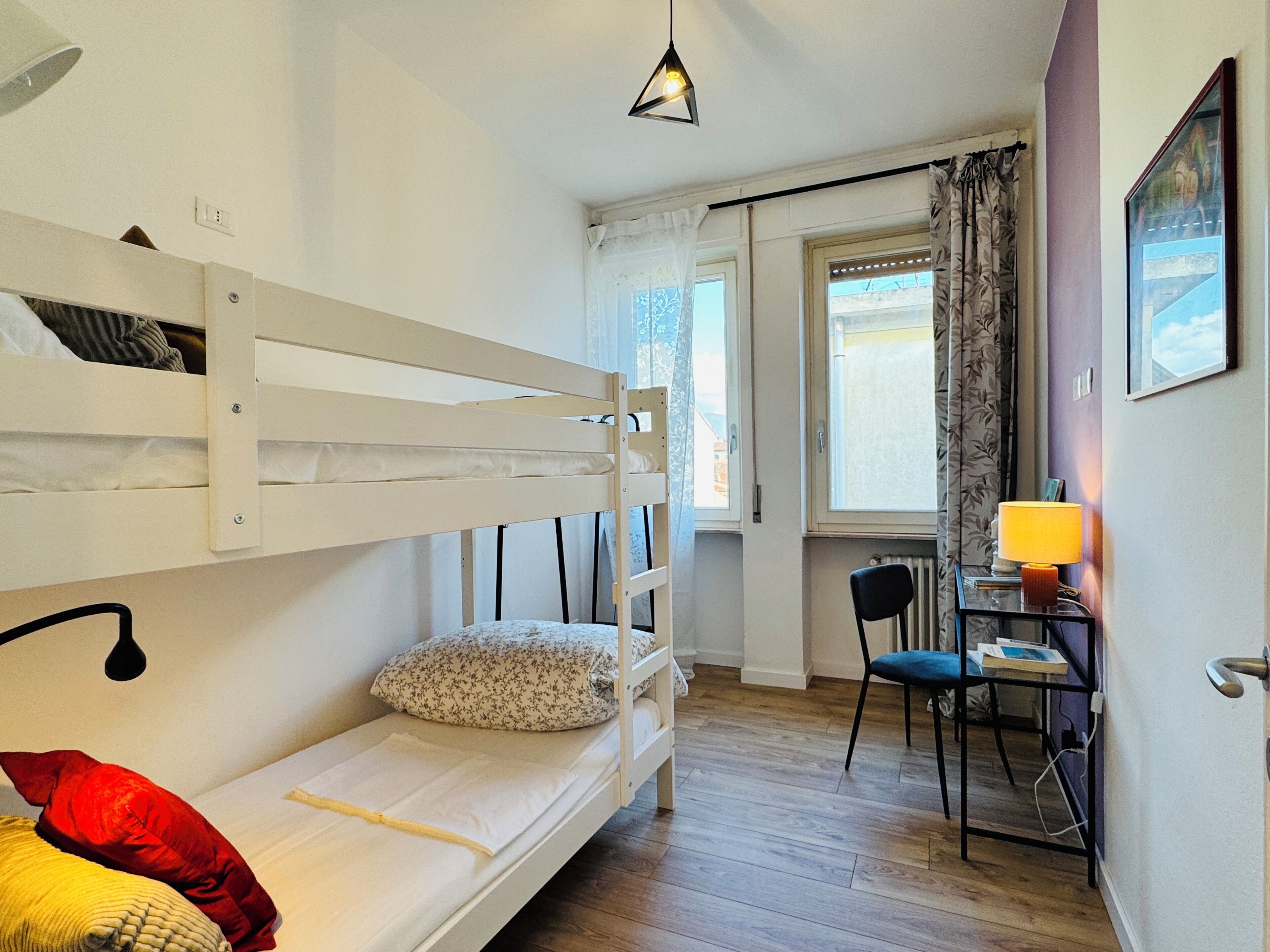 A bunk bed with a pillow, a desk with a chair, a lamp, and a window with curtains.