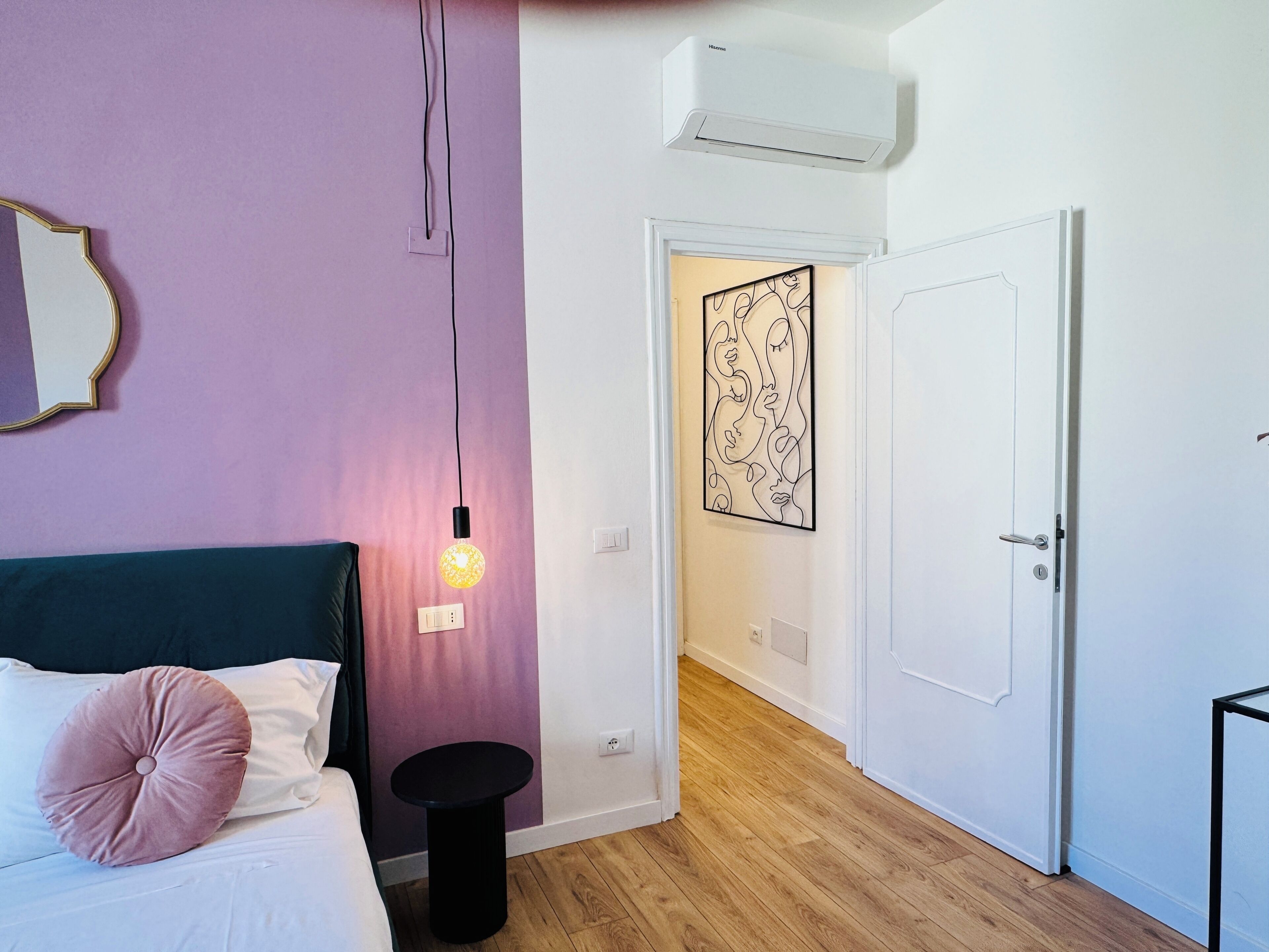 A bedroom with a bed, a bedside table, a wall-mounted air conditioner, and a door leading to another room.