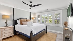 Ein Schlafzimmer mit einem Bett, Nachttischen, einem Deckenventilator, einem Fenster mit Jalousien und einem am Wand montierten Fernseher.