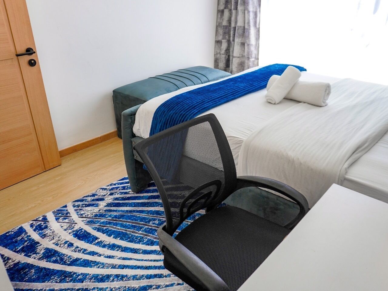 Hotellihuone, jossa on sänky, työpöytä tuolilla, sinivalkoinen kuvioitu matto ja puinen ovi.