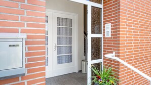 A brick building with a white door, a metal trash can, and a potted plant with flowers.