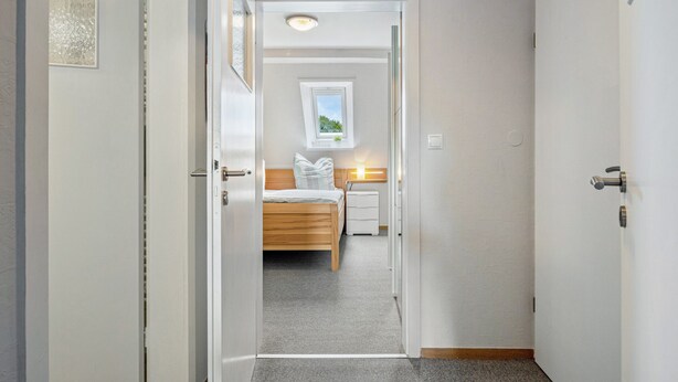A narrow hallway leading to a bedroom with a bed, bedside table, and a window.
