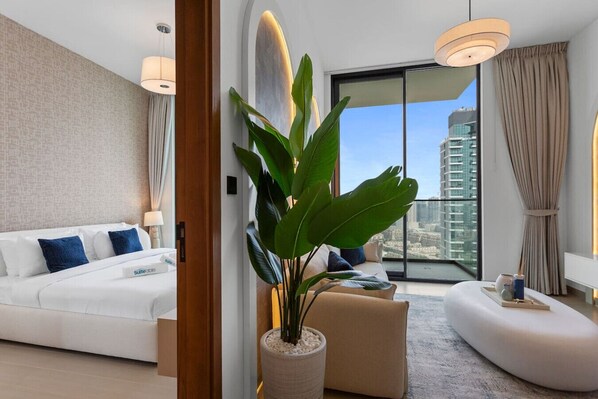 A modern hotel room with a white sofa, a large potted plant, a round seating area, and a view of a cityscape through large windows.